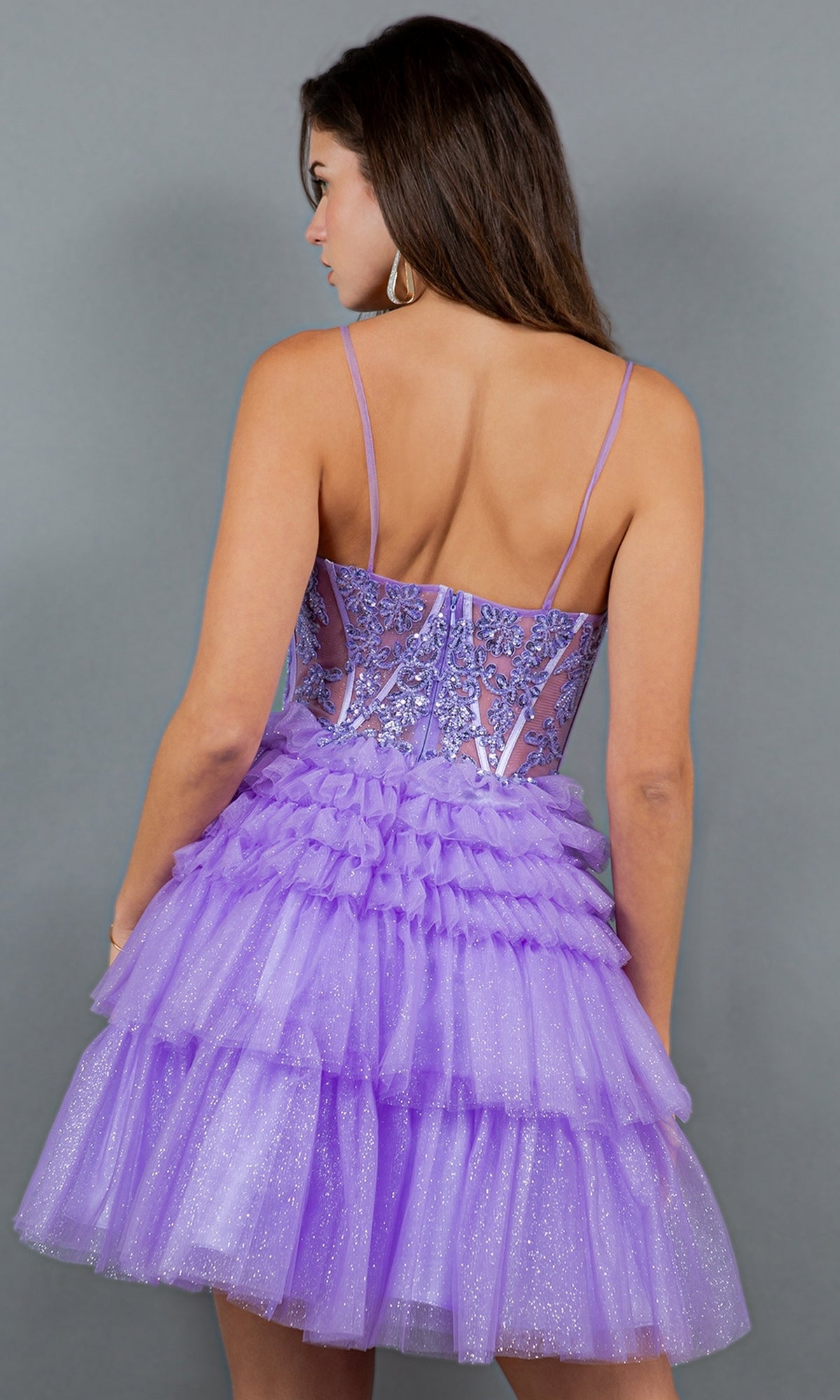 A woman with long brown hair wears the Short Sweetheart Ruffled Homecoming Dress 5133J, a lilac A-line dress with a sheer embroidered bodice and layered tulle skirt, while facing away against a gray background.
